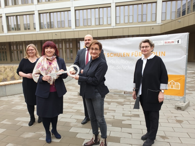 Mehrere Menschen stehen vor einem Schulgebäude und einem Plakat mit der Aufschrift 